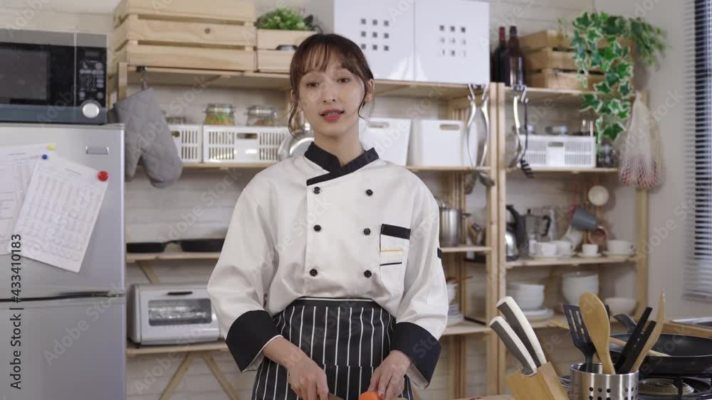 female chef recording video for her channel at home kitchen while prepping for food with knife. looking at camera, asian woman vlogger sharing cooking tips on social media with hand gesture.