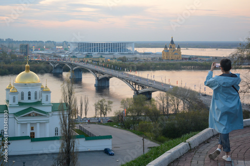 NIZHNY NOVGOROD, RUSSIA - MAY 6, 2021: Girl is taking a photo of Strelka, cathedral and stadium from Fedorovsky emankment © Andrey