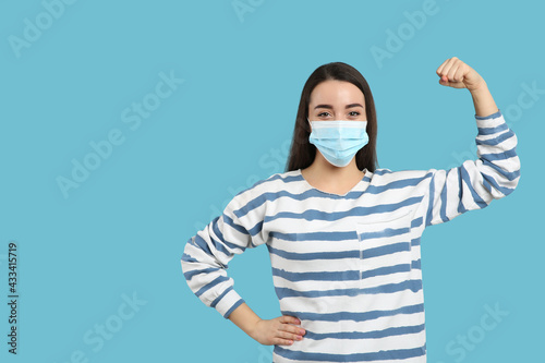 Woman with protective mask showing muscles on light blue background, space for text. Strong immunity concept