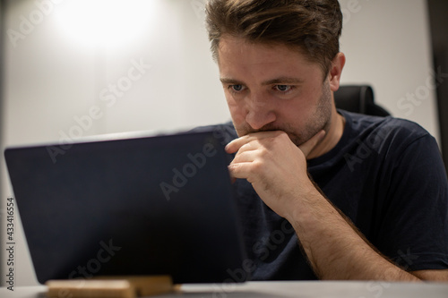 Close up portrait of Focused unshaven handsome guy looking at tablet screen, working from home remotely, participating in online conference at white interior. Male staying connected during pandemic