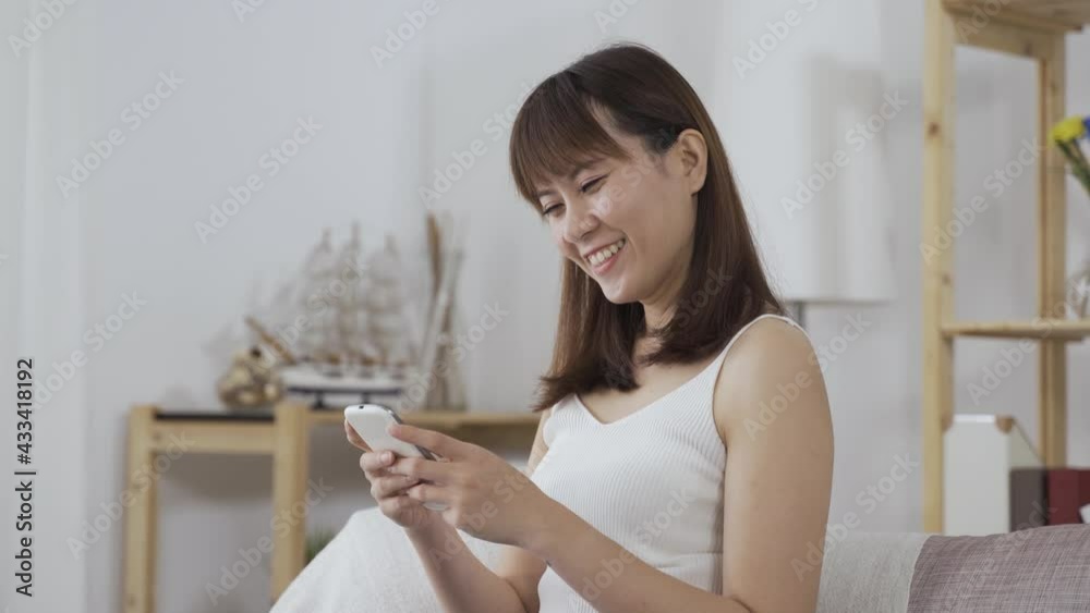 pretty Asian woman having a hearty laugh, sitting straight on the sofa is texting her friends with chatting app on phone. pretty Asian woman having a hearty laugh, sitting straight on the sofa is texting her friends with chatting app on phone.