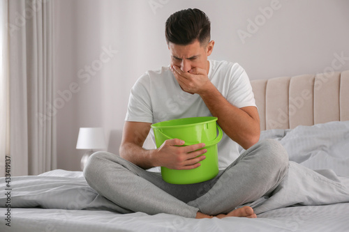 Man with bucket suffering from nausea on bed at home. Food poisoning photo