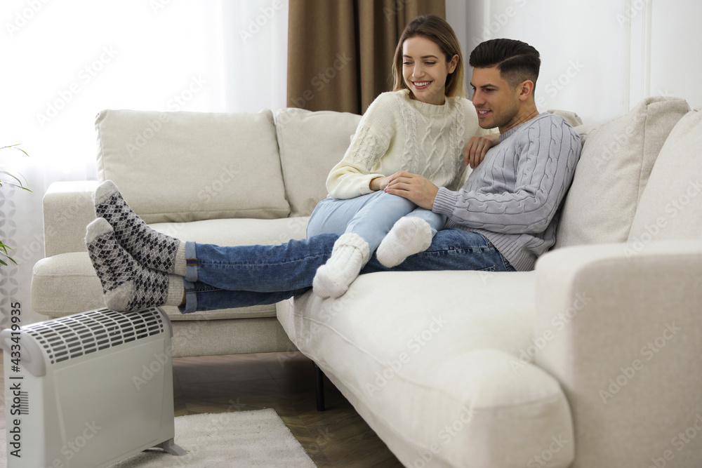 Happy couple sitting on sofa near electric heater at home Happy couple sitting on sofa near electric heater at home