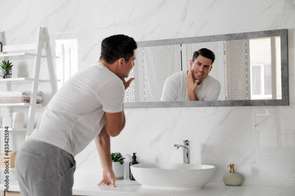 Fototapeta premium Handsome man touching his smooth face after shaving near mirror in bathroom