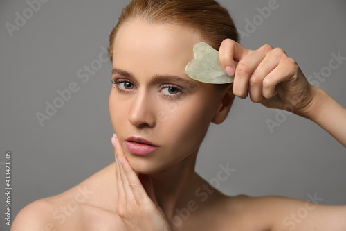 Beautiful young woman doing facial massage with gua sha tool on grey background, closeup