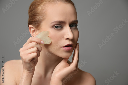 Beautiful young woman doing facial massage with gua sha tool on grey background, closeup