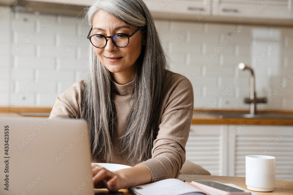 Mature grey woman working with laptop while sitting in kitchen at home Mature grey woman working with laptop while sitting in kitchen at home