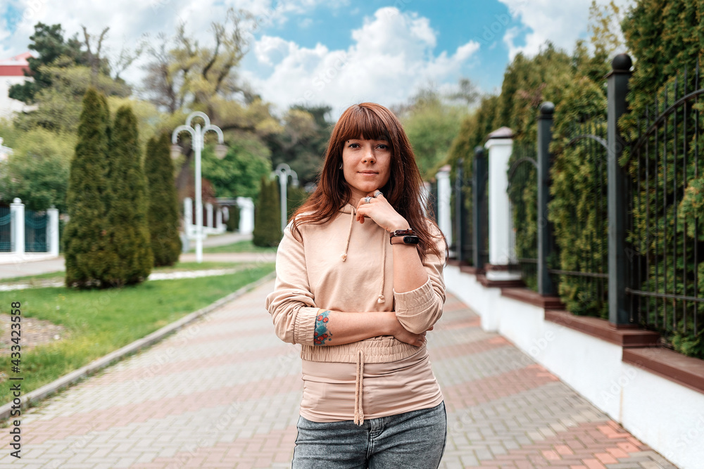 Portrait of a young, confident woman with her tattoed arms posing at the street. In the background is an park. The concept of successful people