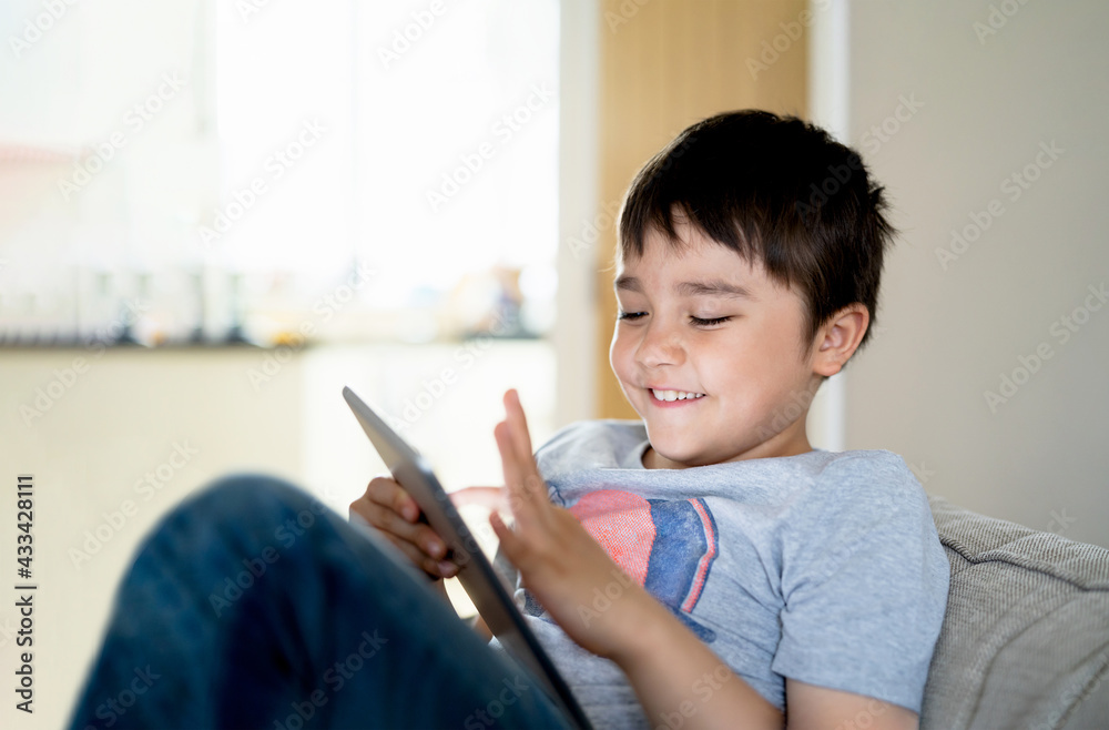 Authentic Kid sitting on sofa watching cartoons or playing games on tablet,Child boy using digital pad learning lesson on internet,Home schooling,Distance learning online education concept Authentic Kid sitting on sofa watching cartoons or playing games on tablet,Child boy using digital pad learning lesson on internet,Home schooling,Distance learning online education concept