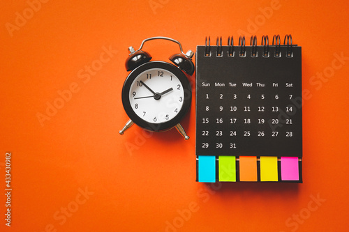 black alarm clock and opened calendar with multi color paper tag   on grunge orange paper background, top view