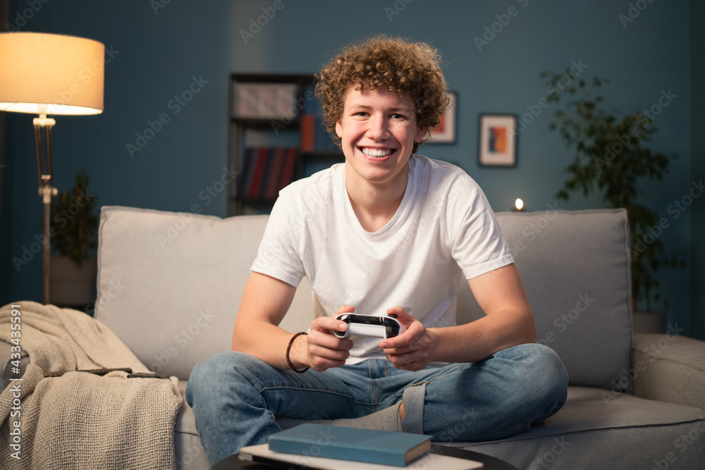 Portrait of smiling young boy, student with curly hair who is looking at the camera. The teenager is pressing buttons from a pad, playing on a console. Portrait of smiling young boy, student with curly hair who is looking at the camera. The teenager is pressing buttons from a pad, playing on a console.