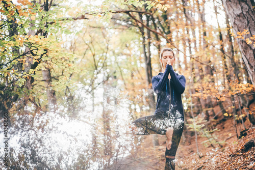 Pretty tranquil woman with ponytail in blue contemporary tracksuit meditates doing namaste mudra among colorful autumn forest on nice sunny day