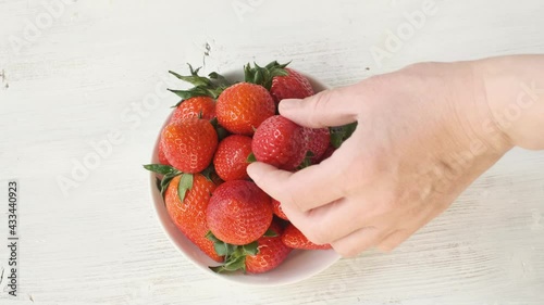 A hand placing a strawberry in a plate. Beautifully stacked red strawberries on a white wooden background.
