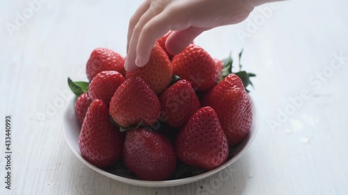 a child's hand taking a strawberry from a plate. Beautifully stacked red strawberries on a white wooden background.