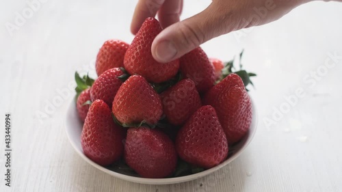 a hand placing a strawberry berry on a plate. Beautifully stacked red strawberries on a white wooden background.