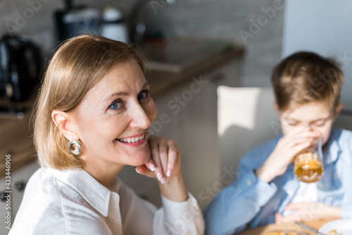 Woman in kitchen looks at the camera, against background of a child drinks juice