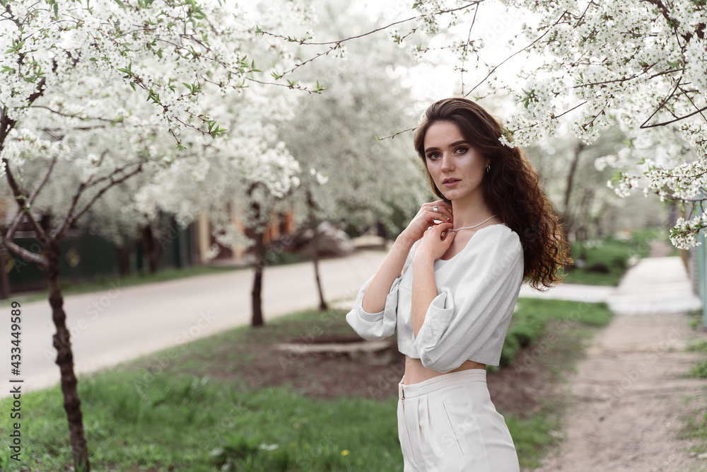 Spring lifestyle. Model posing near white blooming trees without mask outdoor countryside. Dreaming girl with curly hair. Spring lifestyle. Model posing near white blooming trees without mask outdoor countryside. Dreaming girl with curly hair.