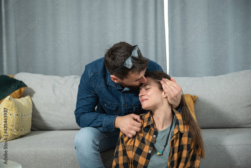Fototapeta premium Young gentle man kissing his girlfriend in the forehead while she sitting on the floor and he on the sofa in their cozy apartment
