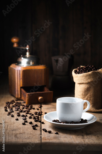 The white coffee cup on the dish. top the old wood table has coffee vintage grinder beans and Moka pot as dark tone background the Top copy space