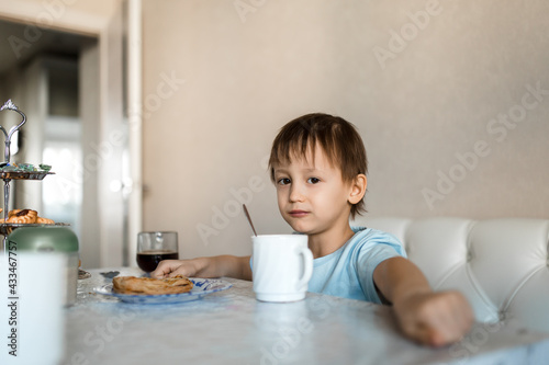 Child sits at the kitchen table and looks at camera, cup and sweets are on table