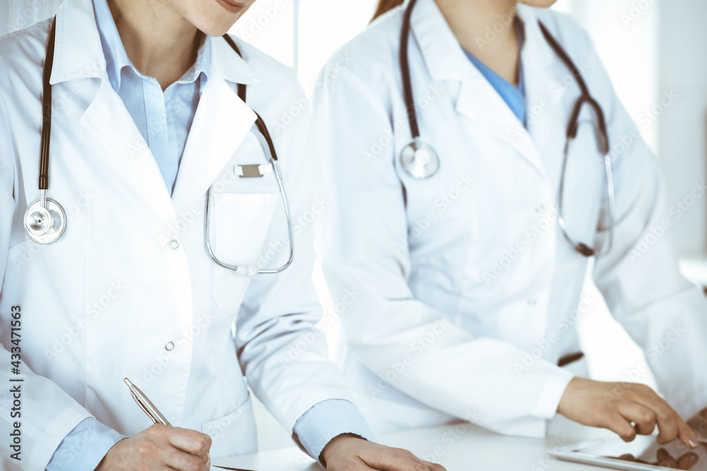 Two woman-doctors at work with patient at background. Female physicians filling up medical documents or prescription while standing in hospital reception desk. Data in medicine Two woman-doctors at work with patient at background. Female physicians filling up medical documents or prescription while standing in hospital reception desk. Data in medicine