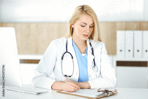 Woman-doctor at work while sitting at the desk in hospital or clinic. Blonde cheerful physician filling up medication history record form