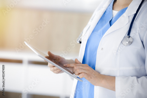 Unknown woman-doctor standing in sunny clinic and using tablet pc, close-up. Data and best service in medicine