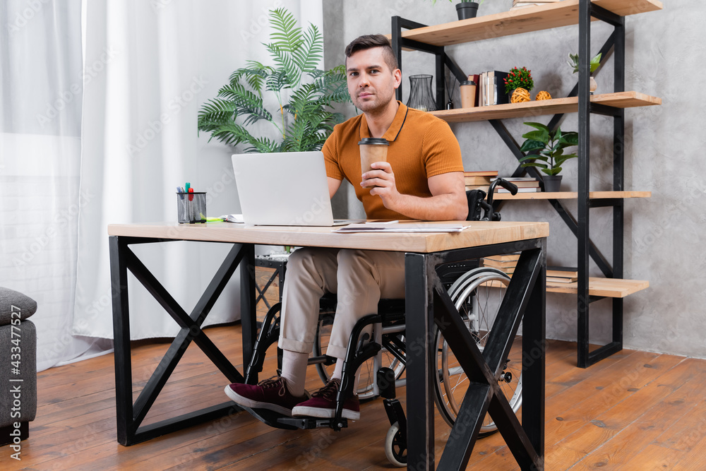 disabled man holding coffee to go while working near laptop at home disabled man holding coffee to go while working near laptop at home