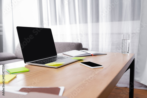 laptop and smartphone with blank screen near glass of water, sticky notes and notebook on desk