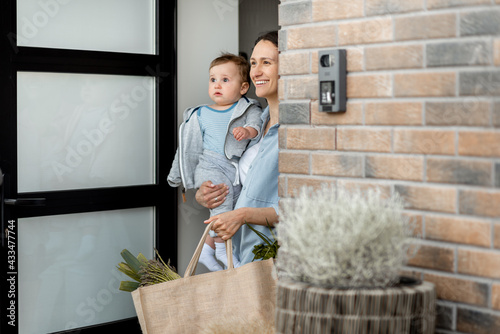 Happy housewife with newborn son staying in front of door of house and bring a shopping bag with products from courier. Online order of food. photo
