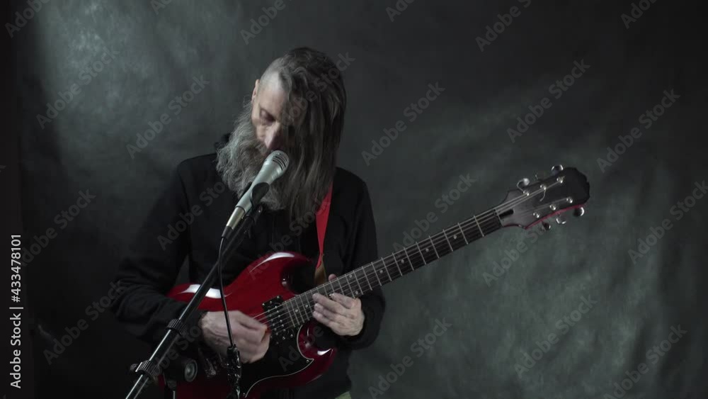 Rockstar with shaved temples, beard and black hoodie plays red electric guitar on stage with black background Rockstar with shaved temples, beard and black hoodie plays red electric guitar on stage with black background