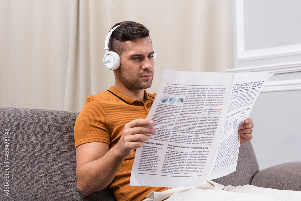 Naklejka premium young man reading newspaper while listening music in wireless headphones