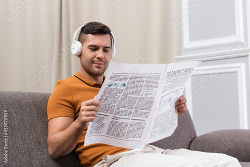 positive man in wireless headphones reading newspaper on sofa at home © LIGHTFIELD STUDIOS