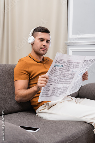 young man reading newspaper and listening music in headphones near smartphone with blank screen