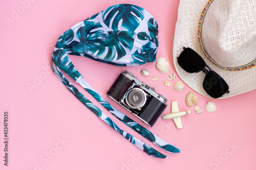 Female straw white hat, swimsuit with palm leaves,black sunglasses, old vintage retro camera, toy plane and seashells isolated on a light color pink background. Top view.Summer vacation travel concept