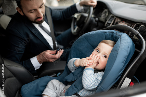 Father tend a child while driving at work with baby sitting in modern car seat. Child new born traveling safety on the road. Safe way to travel fastened seat belts in a vehicle with young kids. 