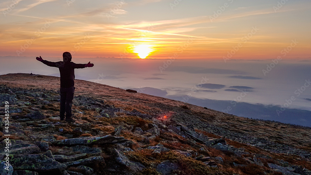 A man in a hoodie standing on top of Babia Gora, Poland, spreading his arms and enjoying the panoramic view on sun rising above the horizon. Thick clouds below. The sky is pink and orange. Freedom