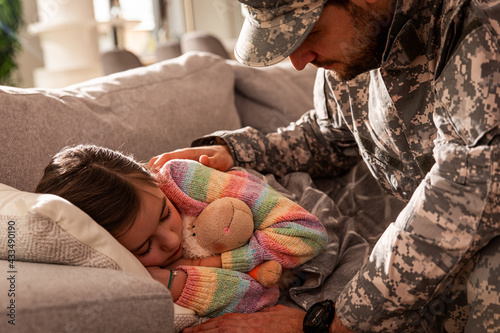 Soldier caresses his daughter while she sleeping on a bed at home.