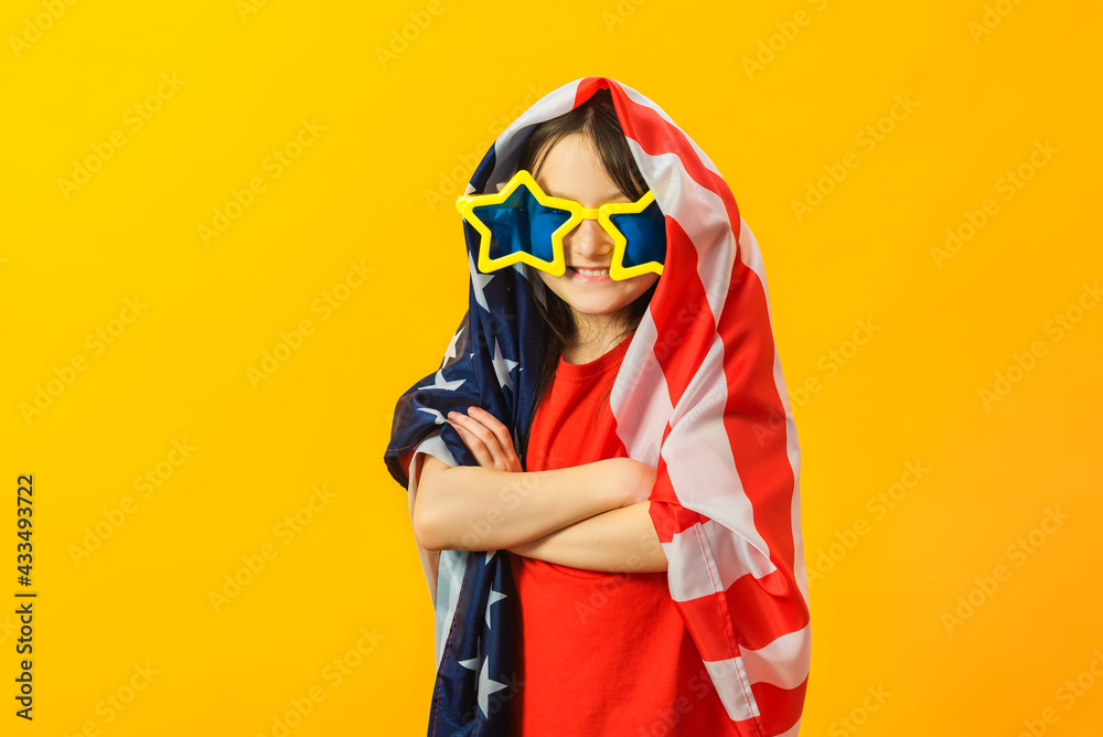 Little pretty girl wrapped in flag of united states of america and posing isolated on yellow studio background. The child wore huge star-shaped sunglasses. Patriot USA concept.