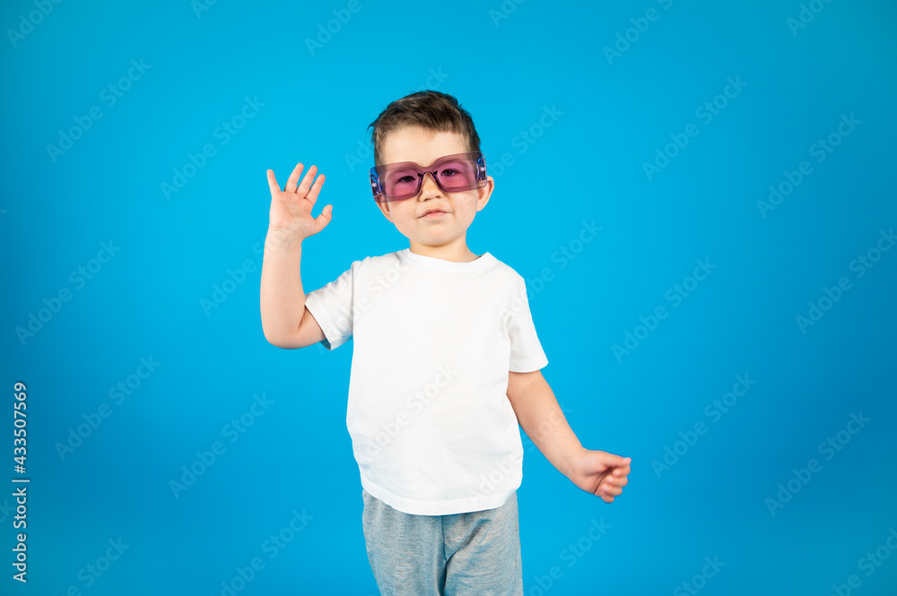 Little funny boy in violet glasses waving into the camera. Blue background with copy space Little funny boy in violet glasses waving into the camera. Blue background with copy space