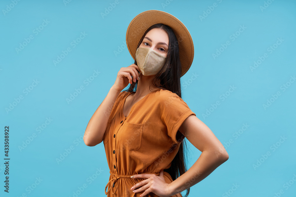 Portrait of beautiful confident young brunette woman wearing cute orange dress, shiny protective mask and boater hat, posing over pastel blue background Portrait of beautiful confident young brunette woman wearing cute orange dress, shiny protective mask and boater hat, posing over pastel blue background