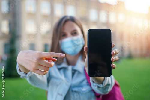  Young smiling student girl in protective medical mask is pointing with index finger at smartphone with blank black screen in her hand