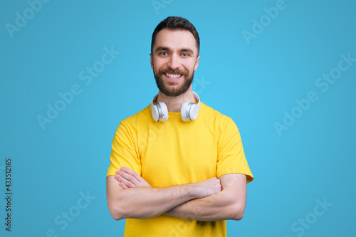 Studio shot of handsome smiling bearded young man wearing basic yellow t-shirt and wireless headset posing over blue background with his hands crossed on the chest