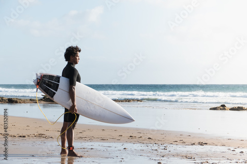Young african malagasy surfer standing at the beach shore watching the waves holding surfboard
