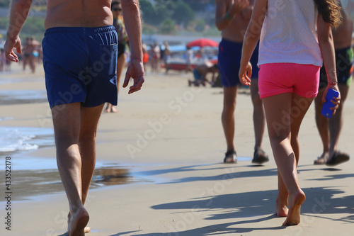 Rear view of a man and a girl walking on an sand beach in the south of Italy. The girl has a fuchsia shorts and a smartphone in the right hand. In the background, out of focus, other swimmers..