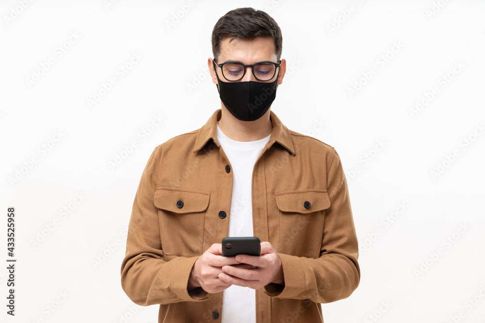 Young modern man in casual brown cargo shirt and black mask, looking at his phone, isolated on gray background. Immunity passport online app Young modern man in casual brown cargo shirt and black mask, looking at his phone, isolated on gray background. Immunity passport online app