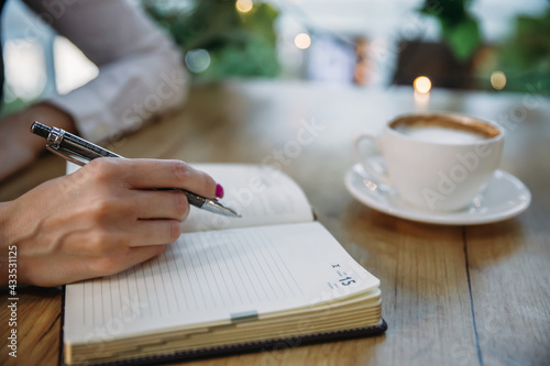 Close-up of a female hand writes in a notebook against the background of a white cup with coffee