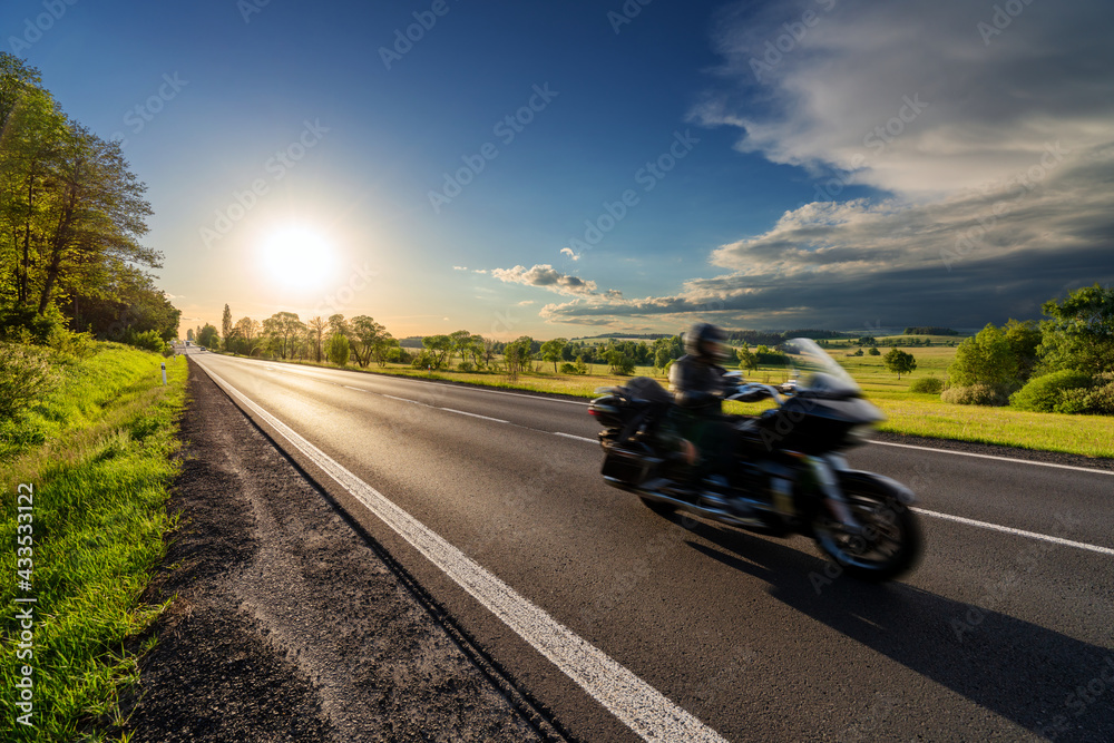 Motion blurred black motorcycle riding on an empty asphalt road in a rural landscape at sunset