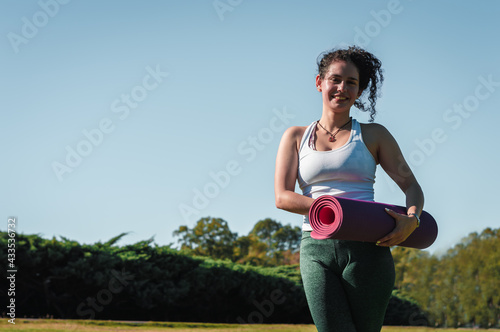 pretty latina girl  with curls  standing in a park smiling with a pink yoga mat in her hands  wearing a green yoga and lifestyle concept.
