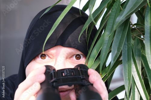 close-up curious islamic woman in war paint, holding field binoculars with zoom, peeping from the bushes, spying on unfaithful husband, neighbors, observation of people and animals photo